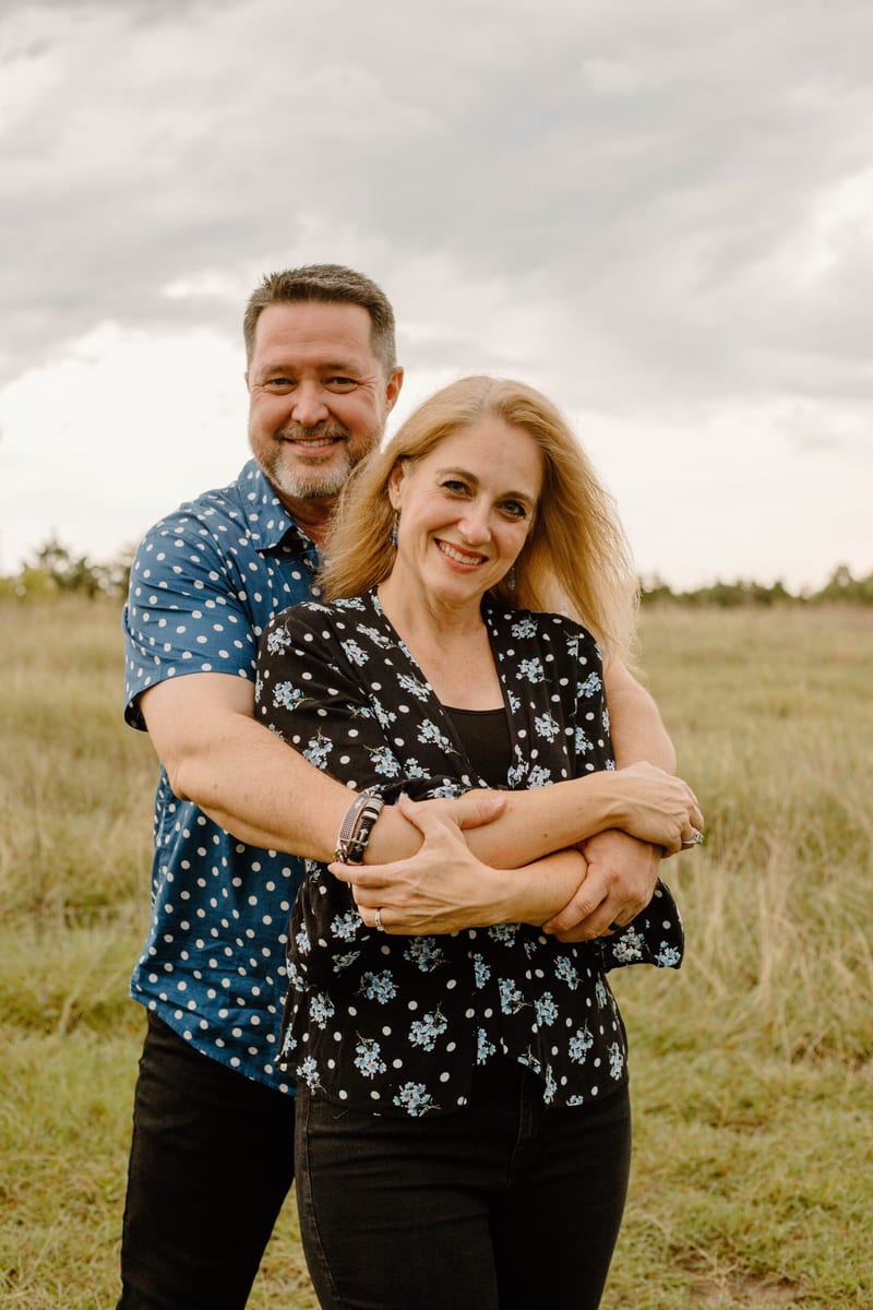 Lafe Angell standing behind his wife Hope with his arms around her in an open field under a cloudy sky.